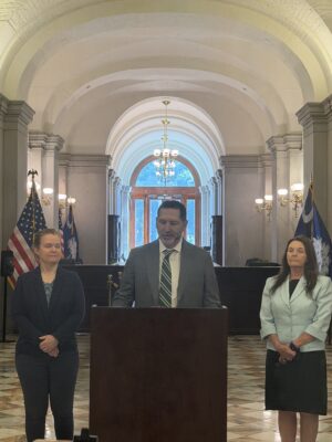 south-carolina-report-card-2 | ASCE's 2025 Infrastructure Report Card Three adults stand behind a wooden podium in a grand arched hallway, a man speaking at center with two women beside him, flags in the background.